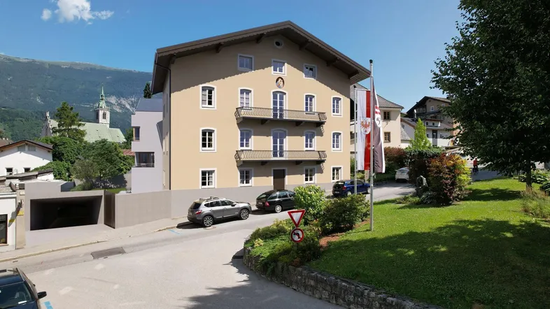Gebäude mit mehreren Balkonen, umgeben von Grünflächen und Bergen im Hintergrund unter blauem Himmel.