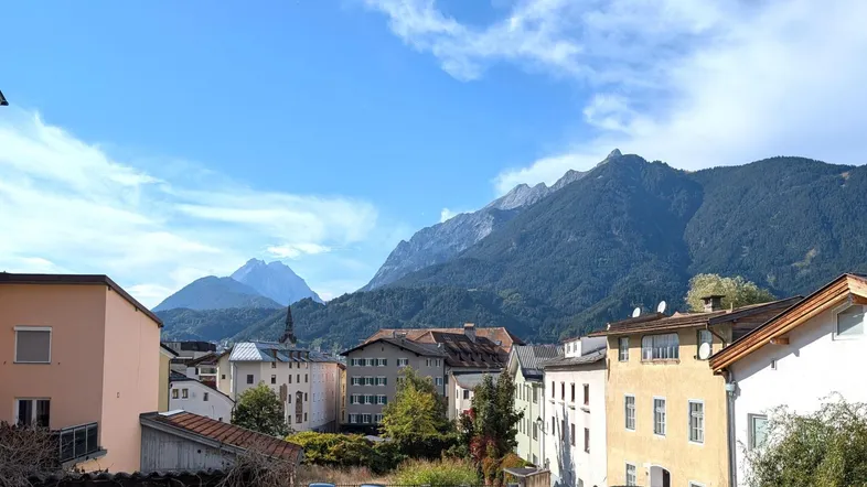Panoramablick über eine Stadt mit traditionellen Gebäuden und majestätischen Bergen im Hintergrund unter blauem Himmel.