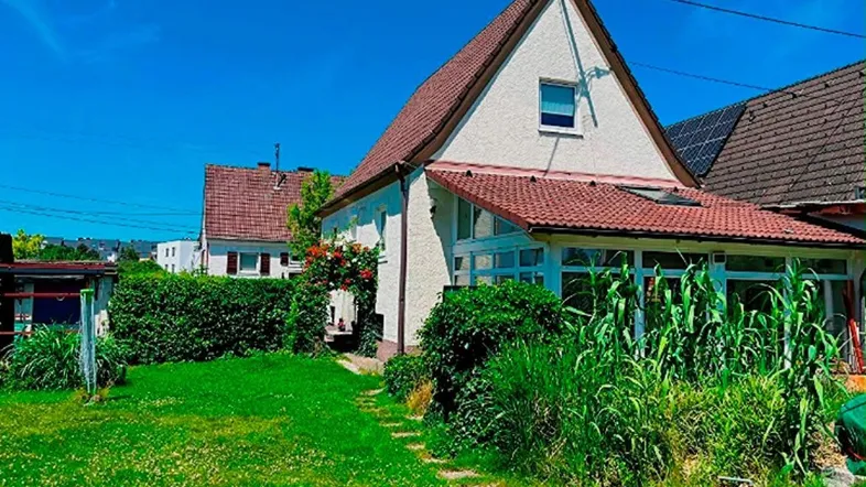 Hausfassade mit grünem Garten, Hecke und einem Wintergarten unter blauem Himmel.