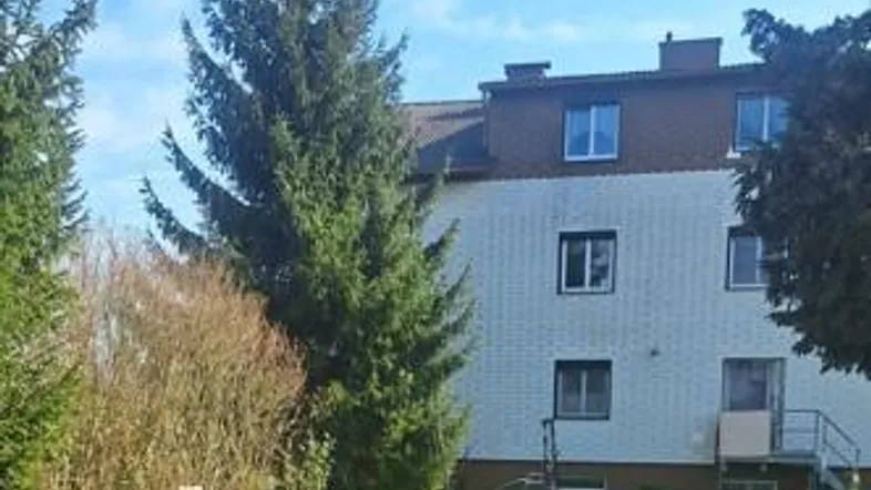 Gebäudefassade mit großem Rasenbereich und hohen Nadelbäumen im Vordergrund unter blauem Himmel.