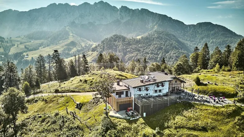 Luftaufnahme eines großen Gebäudekomplexes auf einem grünen Hügel mit majestätischen Bergen im Hintergrund.