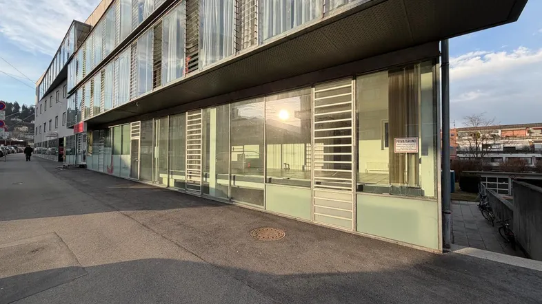 Moderne Geschäftsfassade mit großen Glasfenstern und reflektierendem Sonnenlicht, die eine städtische Umgebung zeigt.