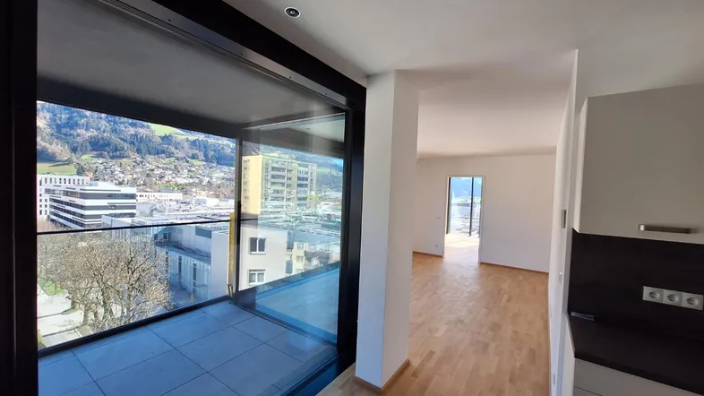 Heller Innenraum mit großen Fenstern, Zugang zum Balkon und Blick auf die Stadt und Berge.
