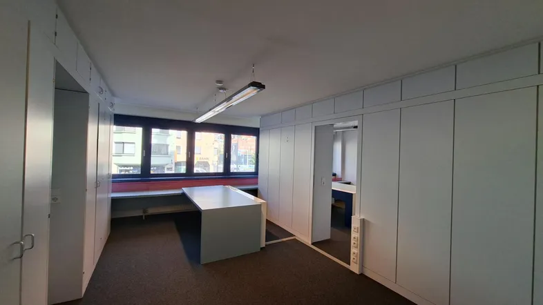 Helles Büro mit großen Fenstern, Einbauschränken und einem zentralen Arbeitstisch.