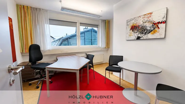 Ein helles Büro mit rotem Teppich, Holzboden und einem Schreibtischbereich mit Blick nach draußen.