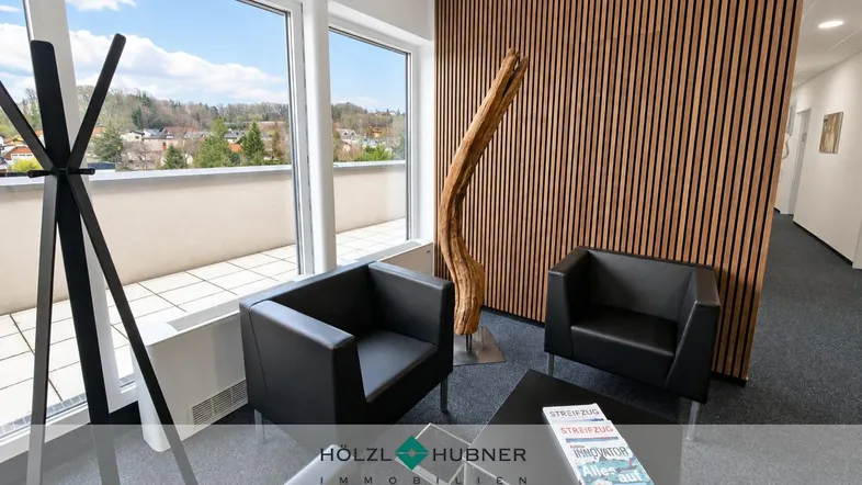 Moderner Empfangsbereich mit bequemen schwarzen Sesseln, Holzwandverkleidung und weitem Ausblick ins Grüne.