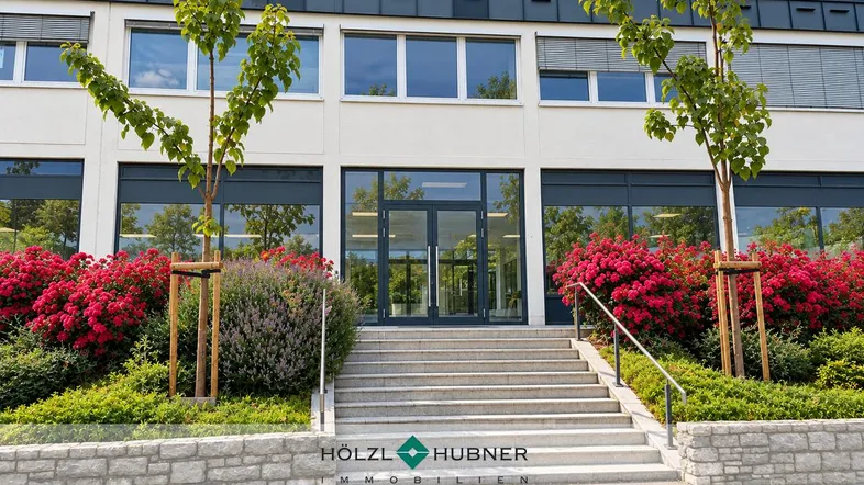 Modernes Gebäude mit großzügigem Eingangsbereich, umgeben von gepflegter Bepflanzung und Bäumen unter blauem Himmel.