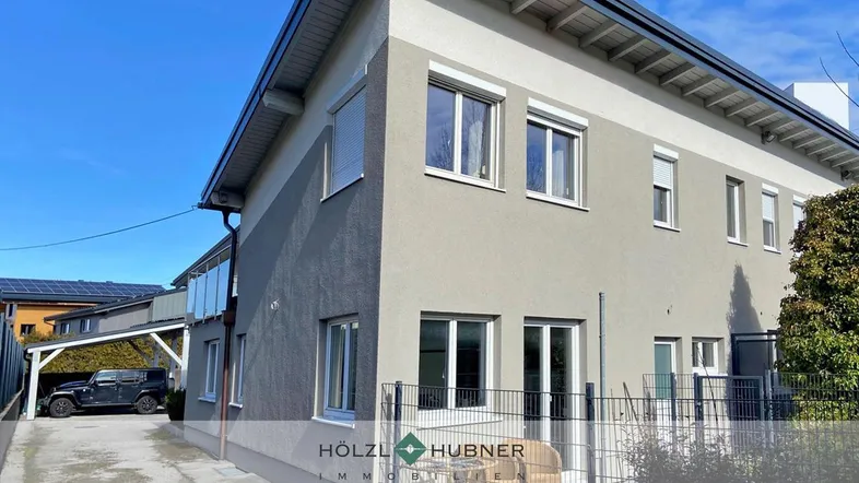 Ansicht des modernen Gebäudes mit einer kleinen Terrasse und einem Carport im Hintergrund unter blauem Himmel.