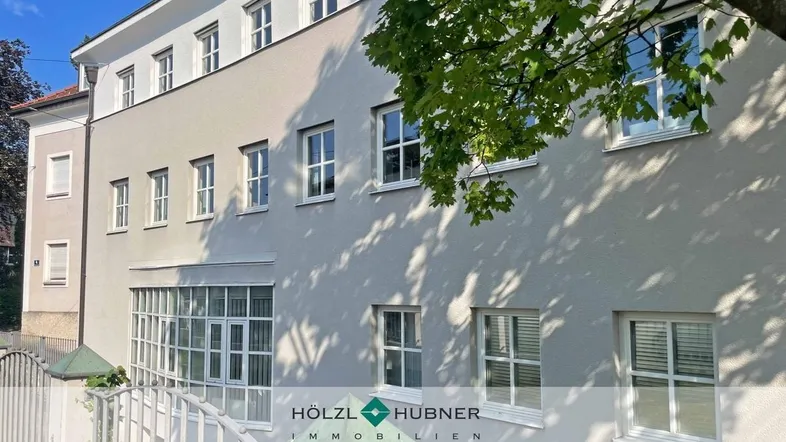 Modernes Gebäude mit heller Fassade und vielen Fenstern, umgeben von grünen Bäumen.