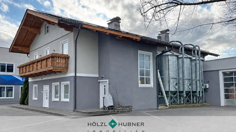 Gebäudeaußenansicht mit traditionellem Holzbalkon, moderner grauer Fassade und angrenzenden Silos.