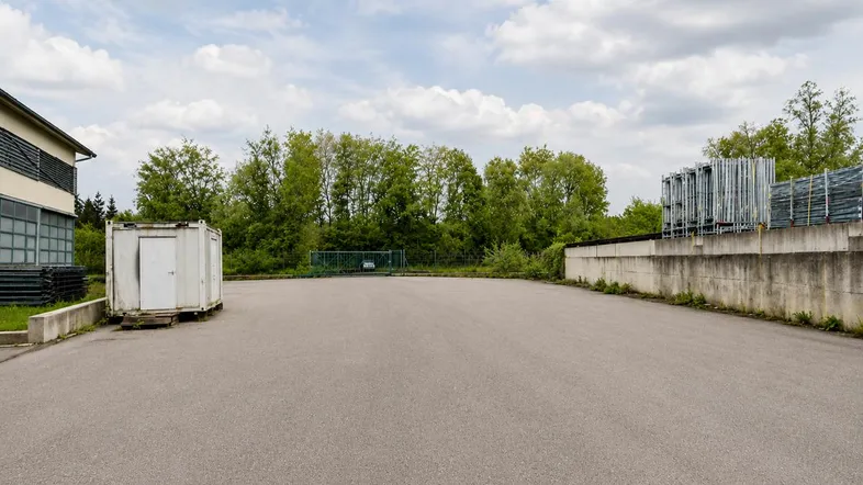 Großer asphaltierter Außenbereich mit einem Gebäude, einem Lagercontainer und Bäumen im Hintergrund unter bewölktem Himmel.