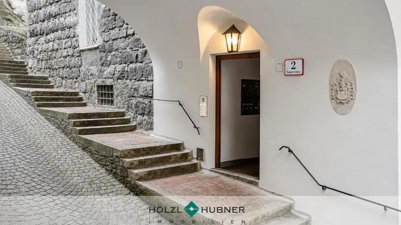 Historischer Eingangsbereich eines Gebäudes mit Steintreppe und gewölbtem Durchgang, der zu einer Haustür führt.