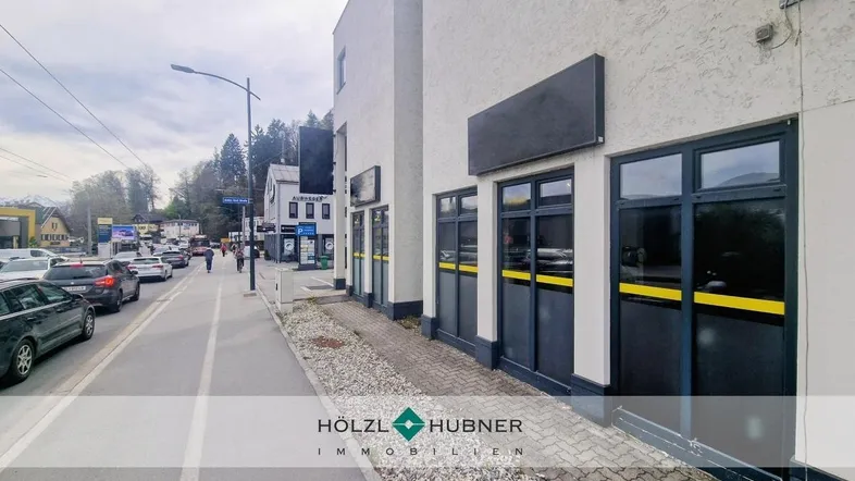 Blick auf die Geschäftsfassade entlang einer belebten Straße mit parkenden Autos und Fußgängern.