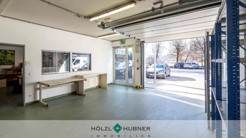 Geräumiger Werkstattbereich mit Arbeitsplatte, Regalen und Blick auf den Außenparkplatz durch das Rolltor.