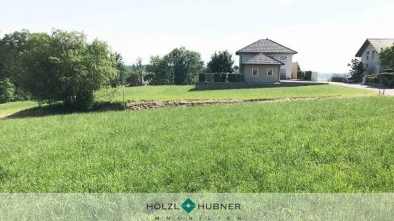 Weitläufige Grünfläche mit Blick auf ein Einfamilienhaus und die umliegende Landschaft unter blauem Himmel.