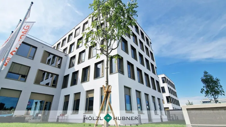 Modernes weißes Bürogebäude mit vielen Fenstern und gepflegter Grünfläche unter blauem Himmel.