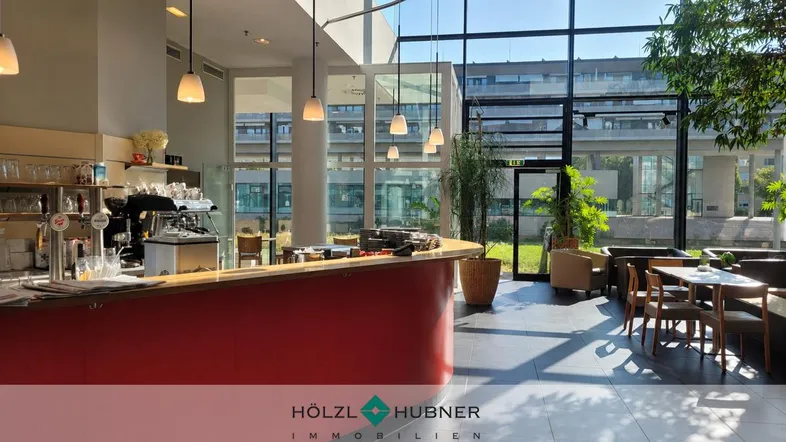 Heller Cafébereich mit roter Theke, großen Glasfronten und Blick ins Grüne.
