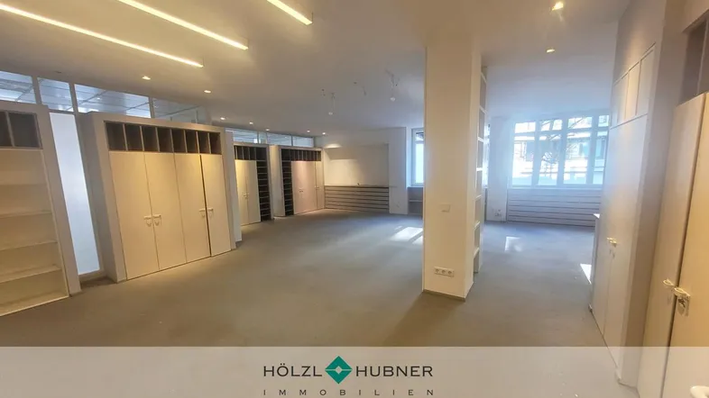 Geräumiger, offener Gewerberaum mit Teppichboden, Einbauschränken und viel Tageslicht durch große Fenster.