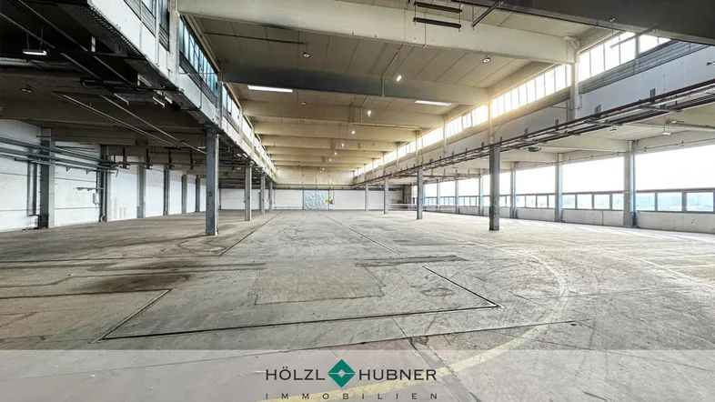 Geräumige, leere Industriehalle mit hohen Decken, Betonboden und großen Fenstern, die viel Tageslicht hereinlassen.