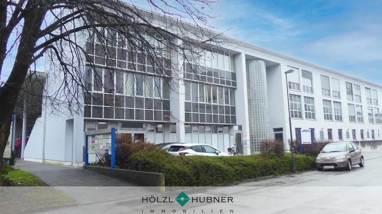 Modernes Bürogebäude mit großen Fensterfronten und Parkplätzen davor unter einem bewölkten Himmel.