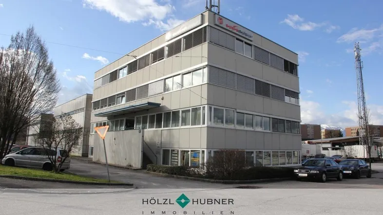 Modernes Bürogebäude mit mehreren Etagen und großen Fenstern unter blauem Himmel und Parkplätzen.