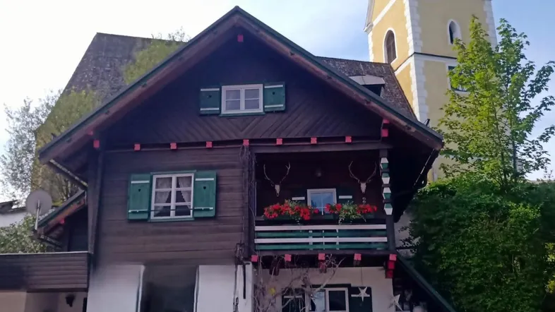 Traditionelles Haus im alpenländischen Stil mit Holzbalkonen und einem Kirchturm im Hintergrund.