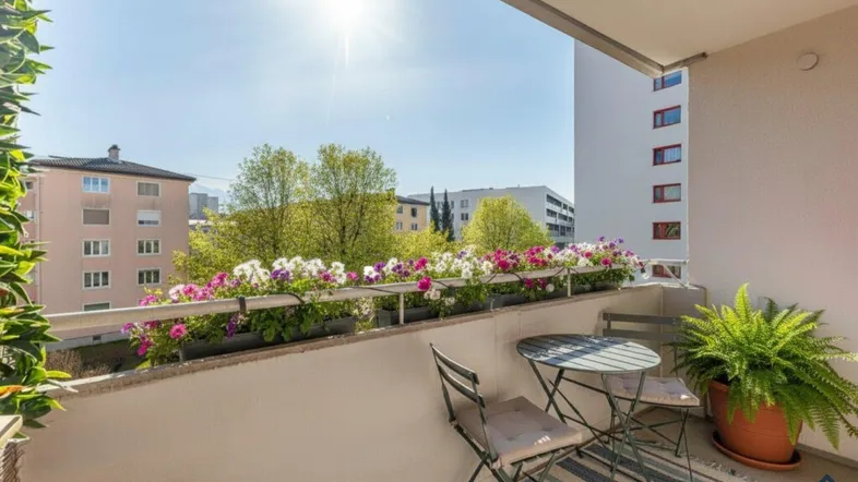 Heller Balkon mit Blumenkästen, Sitzgelegenheiten und Blick auf die umliegenden Gebäude.