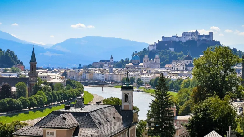 Panoramablick über die historische Stadt Salzburg mit Fluss, Gebäuden und majestätischen Bergen im Hintergrund.