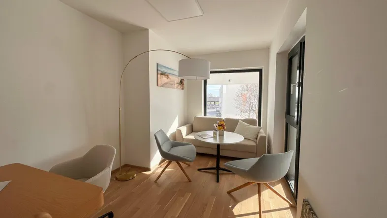 Heller und modern eingerichteter Wohnbereich mit beigem Sofa, zwei Sesseln und Holzboden, durchflutet von Tageslicht.