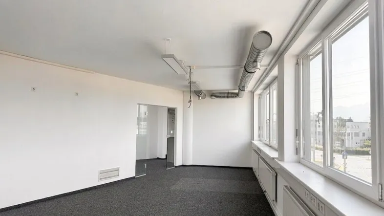 Geräumiges, leeres Büro mit Teppichboden, großen Fenstern und sichtbaren Lüftungsrohren, bietet Bergblick.