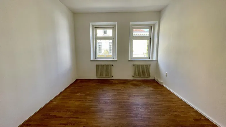 Helles, leeres Zimmer mit Holzboden und zwei Fenstern, bereit zur individuellen Gestaltung.