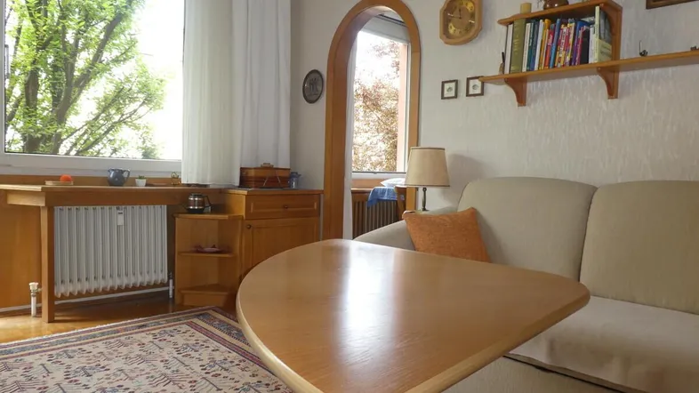 Gemütliches Wohnzimmer mit hellem Sofa, Holztisch und Blick ins Grüne durch große Fenster.