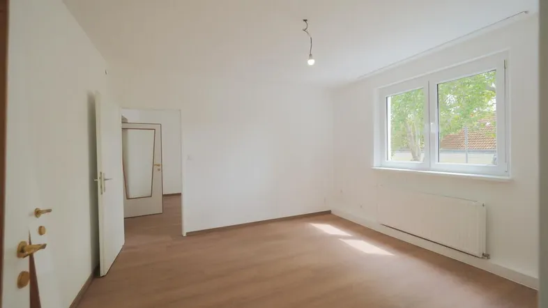 Helles, leeres Zimmer mit Laminatboden, weißen Wänden und einem Fenster, bereit zur individuellen Gestaltung.