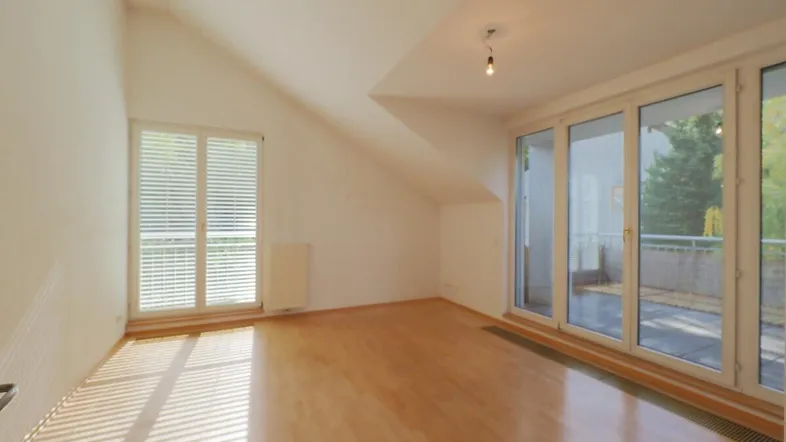 Helles Zimmer mit Holzfußboden, Dachschrägen und großen Glastüren, die zu einem Außenbereich führen.