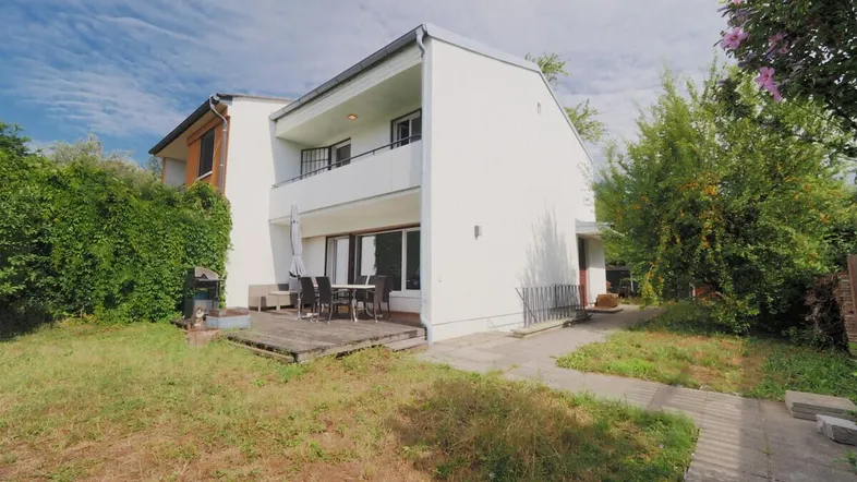 Weißes Haus mit Holzterrasse, Gartenmöbeln und Rasenfläche unter blauem Himmel.
