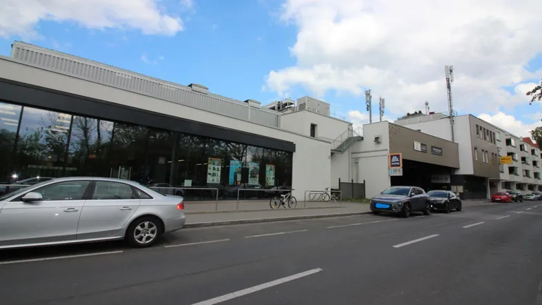 Ansicht eines modernen Geschäftsgebäudes mit Supermarkt und Parkplätzen an einer belebten Straße.