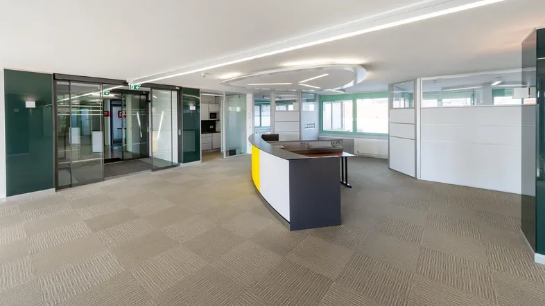 Moderner Empfangsbereich in einem Bürogebäude mit Teppichboden und Glaswänden, hell beleuchtet.