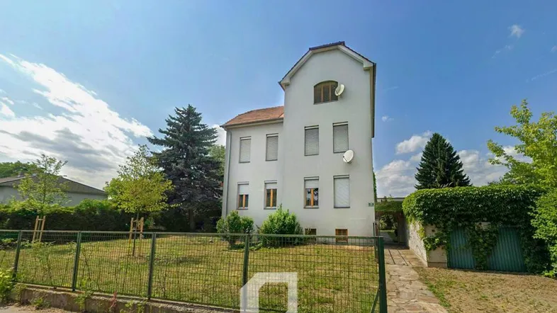 Weißes Gebäude mit Garten und Zaun, umgeben von Bäumen unter blauem Himmel.