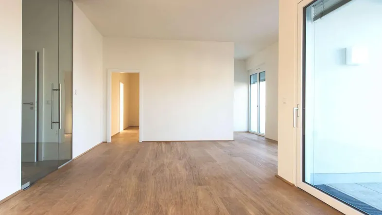 Heller, leerer Raum mit Holzboden und Zugang zu einem Balkon durch eine große Glasfront.