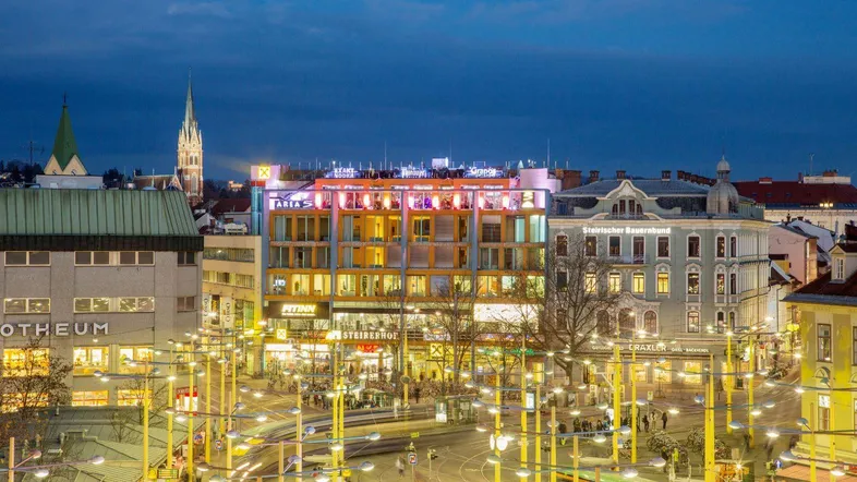 Abendliche Ansicht eines belebten Stadtplatzes mit modernen und historischen Gebäuden unter einem dunklen Himmel.