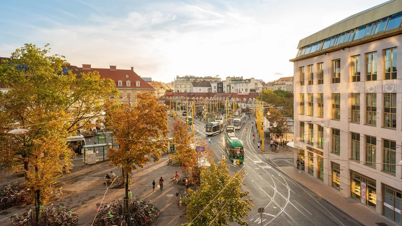 Eine belebte Stadtansicht mit Straßenbahnen, Fahrrädern und Gebäuden unter einem sonnigen Himmel.