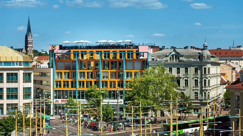 Panoramablick auf eine belebte Stadtstraße mit modernen Gebäuden und historischer Architektur unter blauem Himmel.