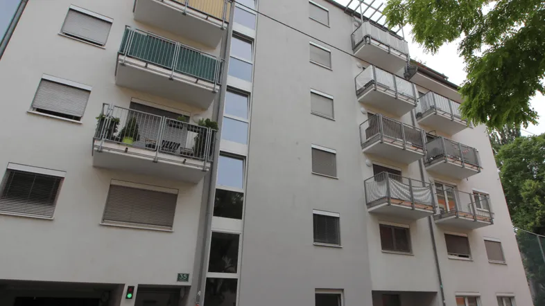 Mehrfamilienhaus mit mehreren Balkonen und Rollläden an den Fenstern, umgeben von Grün.