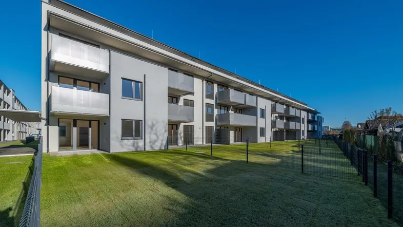 Modernes Mehrfamilienhaus mit Balkonen und Grünflächen, umgeben von einem Zaun.