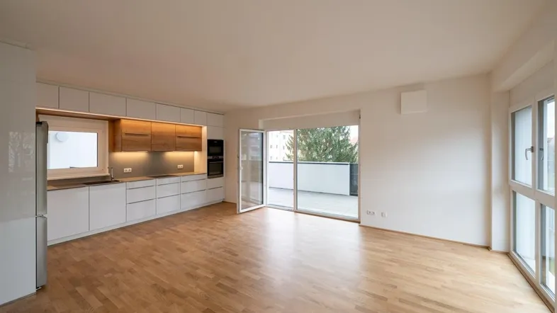 Heller offener Wohnbereich mit moderner Einbauküche, Parkettboden und Zugang zum Balkon.
