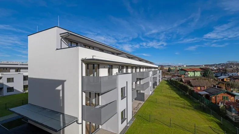 Moderne Wohnanlage mit weißer Fassade, grauen Balkonen und gepflegter Grünfläche unter blauem Himmel.