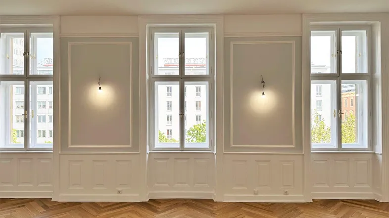 Helles Zimmer mit Fischgrätparkett, hohen Fenstern und eleganter Wandvertäfelung.