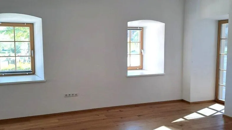 Helles Zimmer mit Holzboden und zwei Fenstern, das viel Tageslicht hereinlässt.