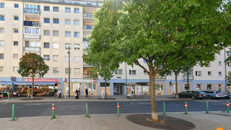 Mehrfamilienhaus mit Geschäften im Erdgeschoss und Bäumen entlang der Straße unter blauem Himmel.