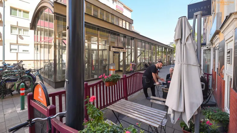 Straßenszene mit Außengastronomie und einem modernen Gebäude mit Glasfassade im Hintergrund.